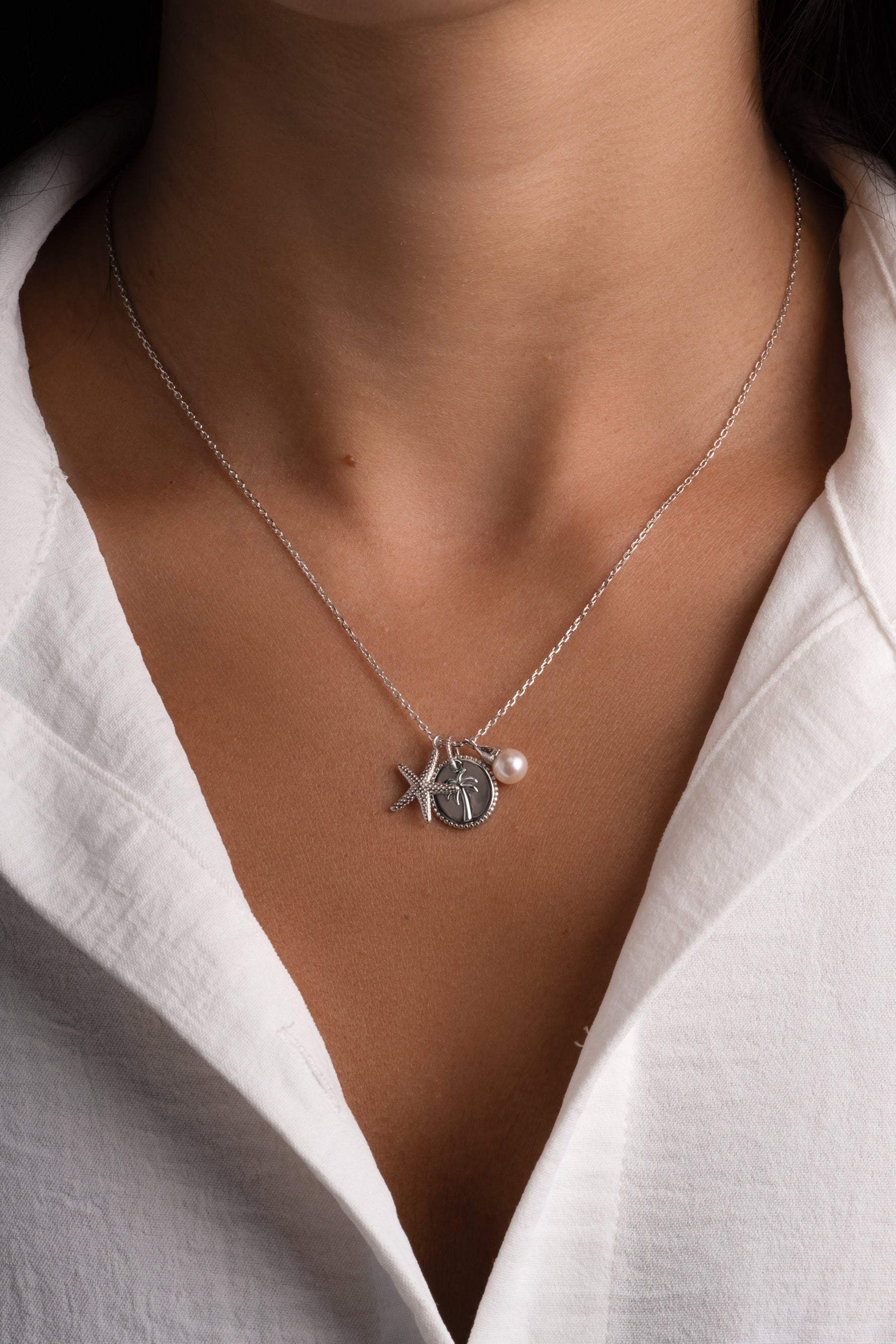 A close-up of a model wearing a delicate silver necklace with a starfish and shell pendant, along with a small pearl accent, styled with a white collared shirt.