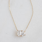 A flat lay of dainty gold necklace, featuring an emerald cut stone set against a pear shaped stone symbolizing two lovers