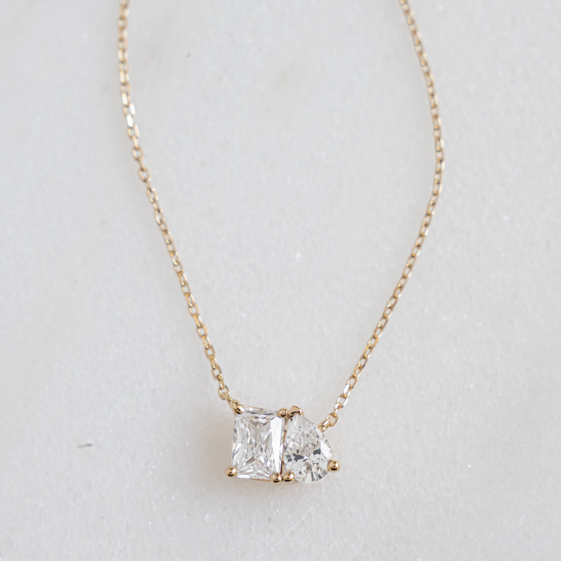 A flat lay of dainty gold necklace, featuring an emerald cut stone set against a pear shaped stone symbolizing two lovers