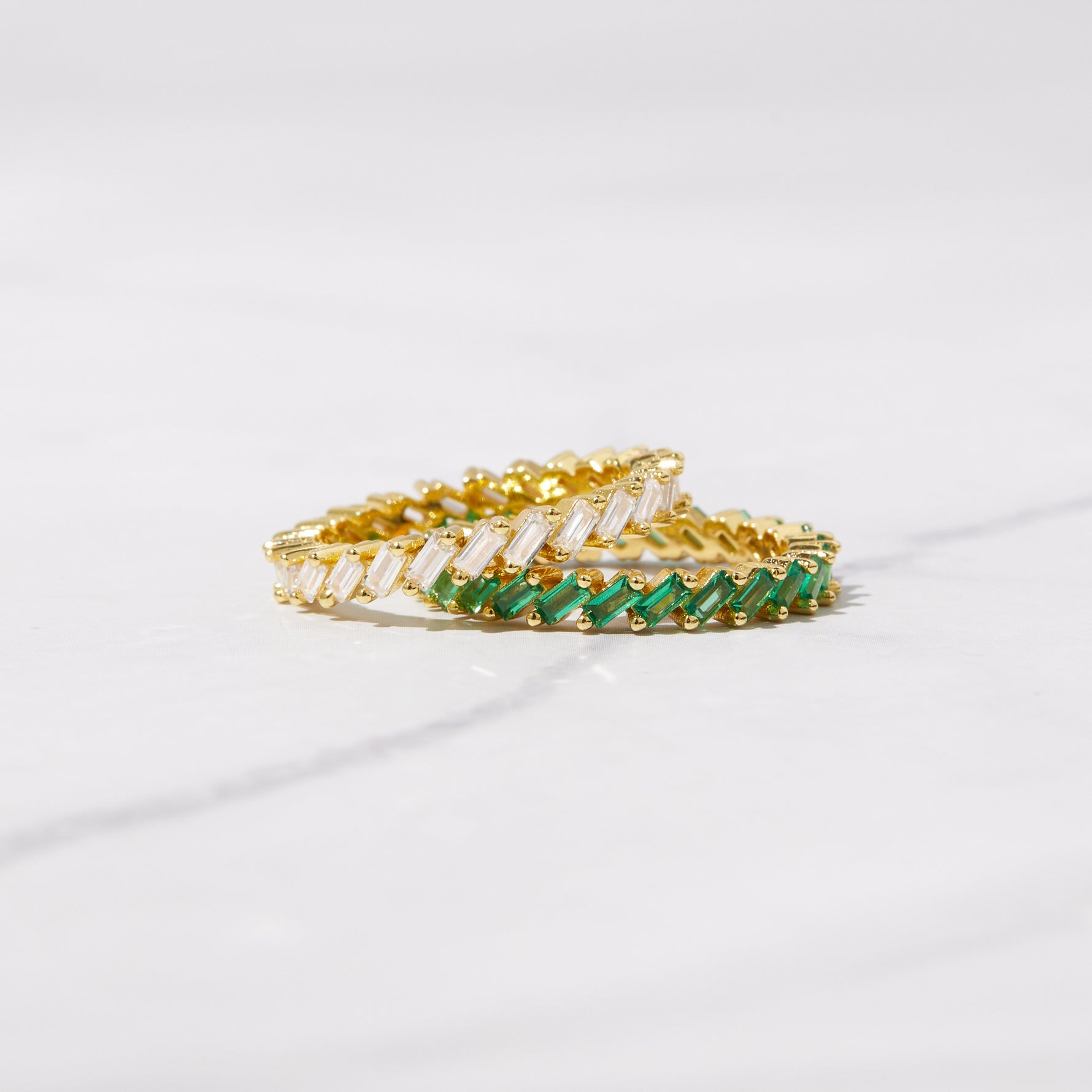 CZ diamond and emerald slanted baguette rings in gold on marble background. 