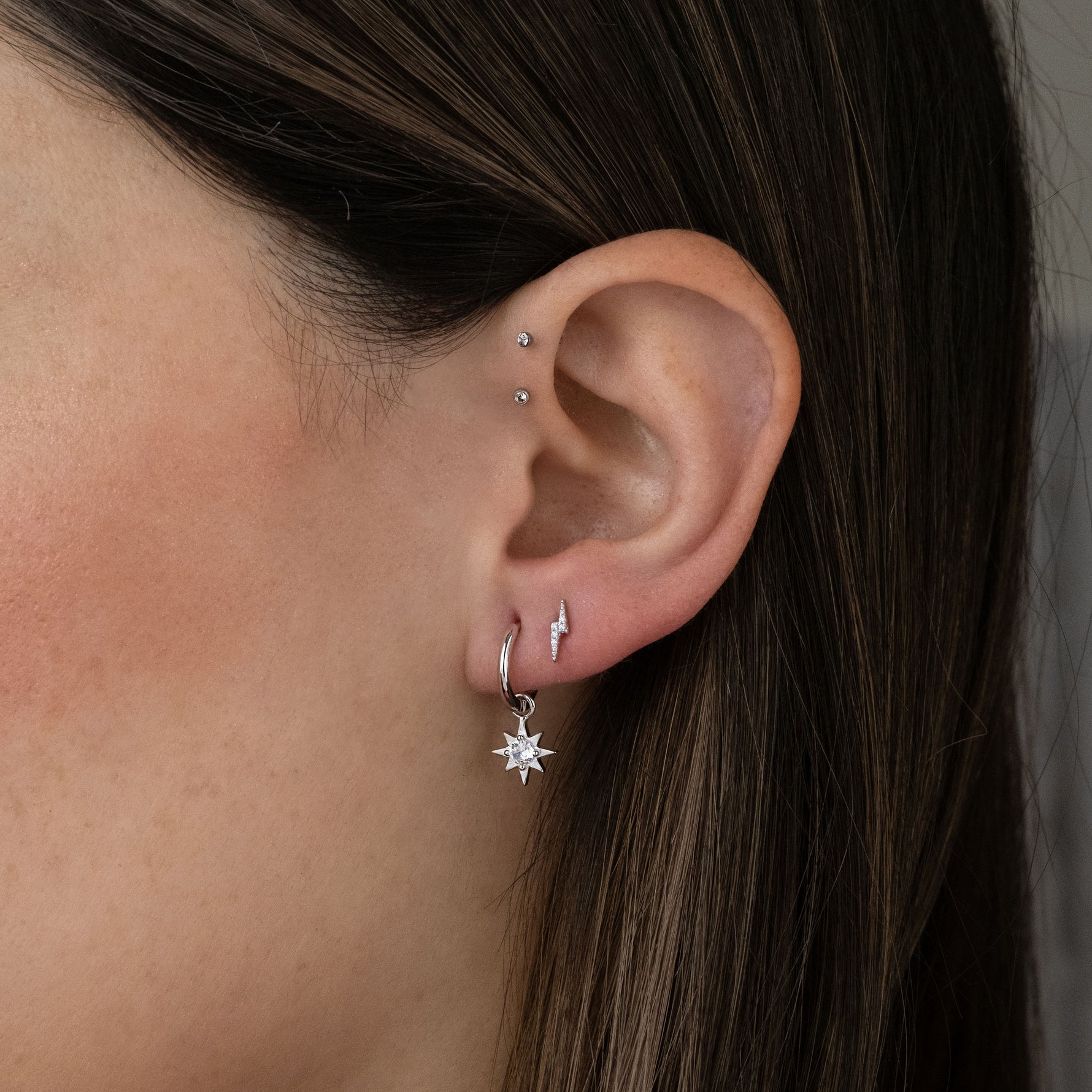 Model wearing a celestial earring stack including a sterling silver sunburst charm huggie and a lightning bolt stud.