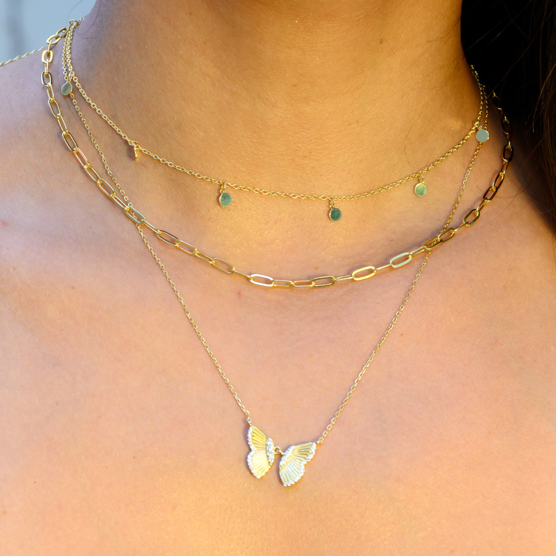 Model wearing delicate gold necklace stack featuring a charm necklace, a paperclip necklace, and a butterfly necklace. 