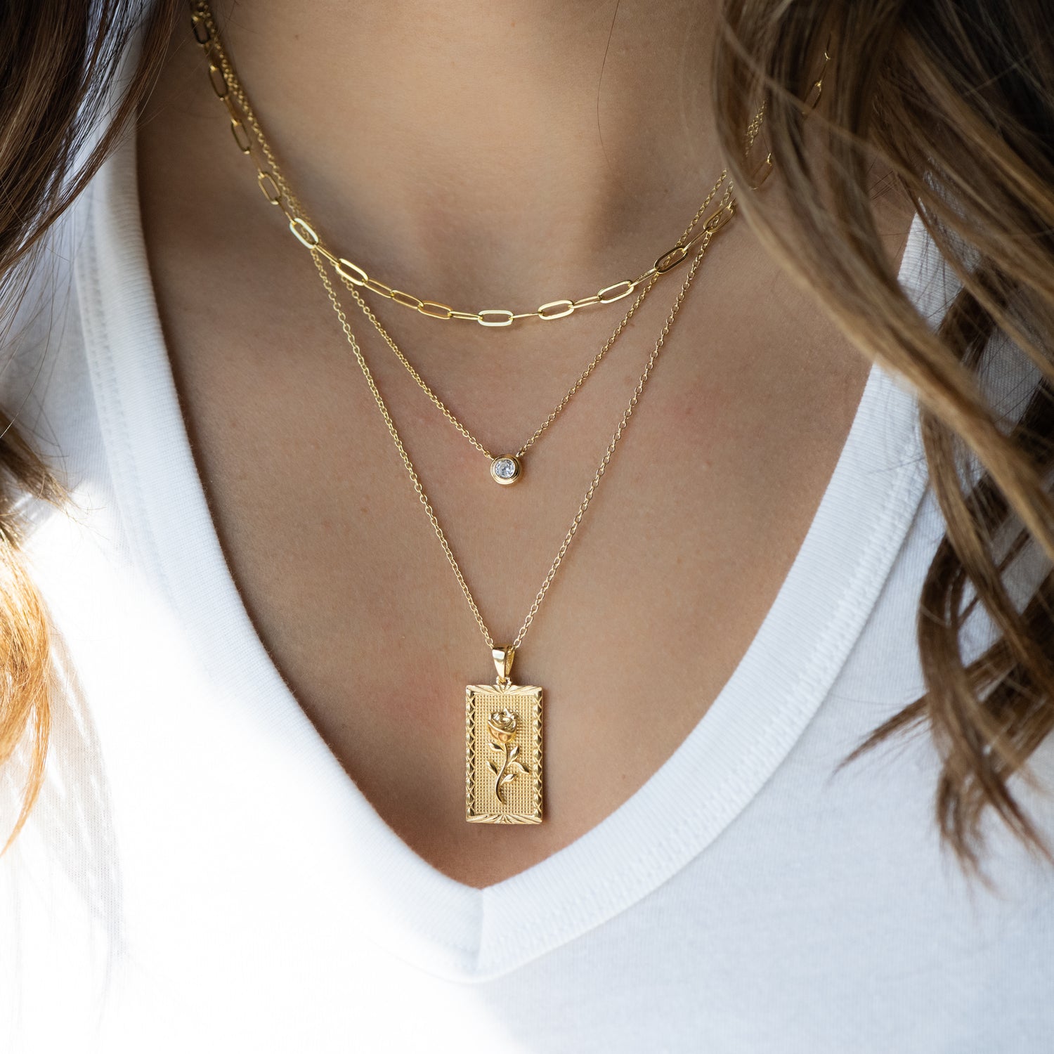 Three layered gold chain necklaces featuring a paperclip necklace, a round solitaire bezel, and a rose rectangle medallion pendant necklace.