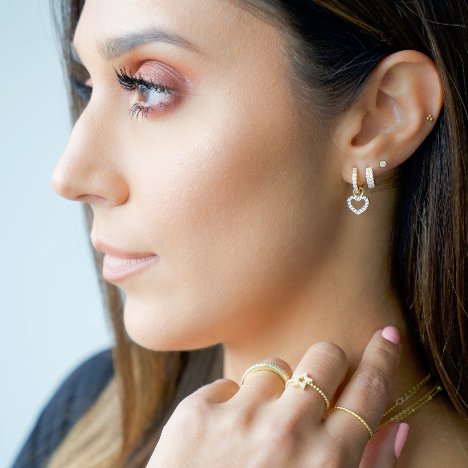 Model wearing pavé feminine gold earring stack including a diamond stud, a diamond huggie, and a heart charm diamond hoop.