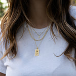 Model wearing three gold necklaces including a tear drop pendant necklace, a square paperclip necklace, and a rose medallion rectangle necklace. 
