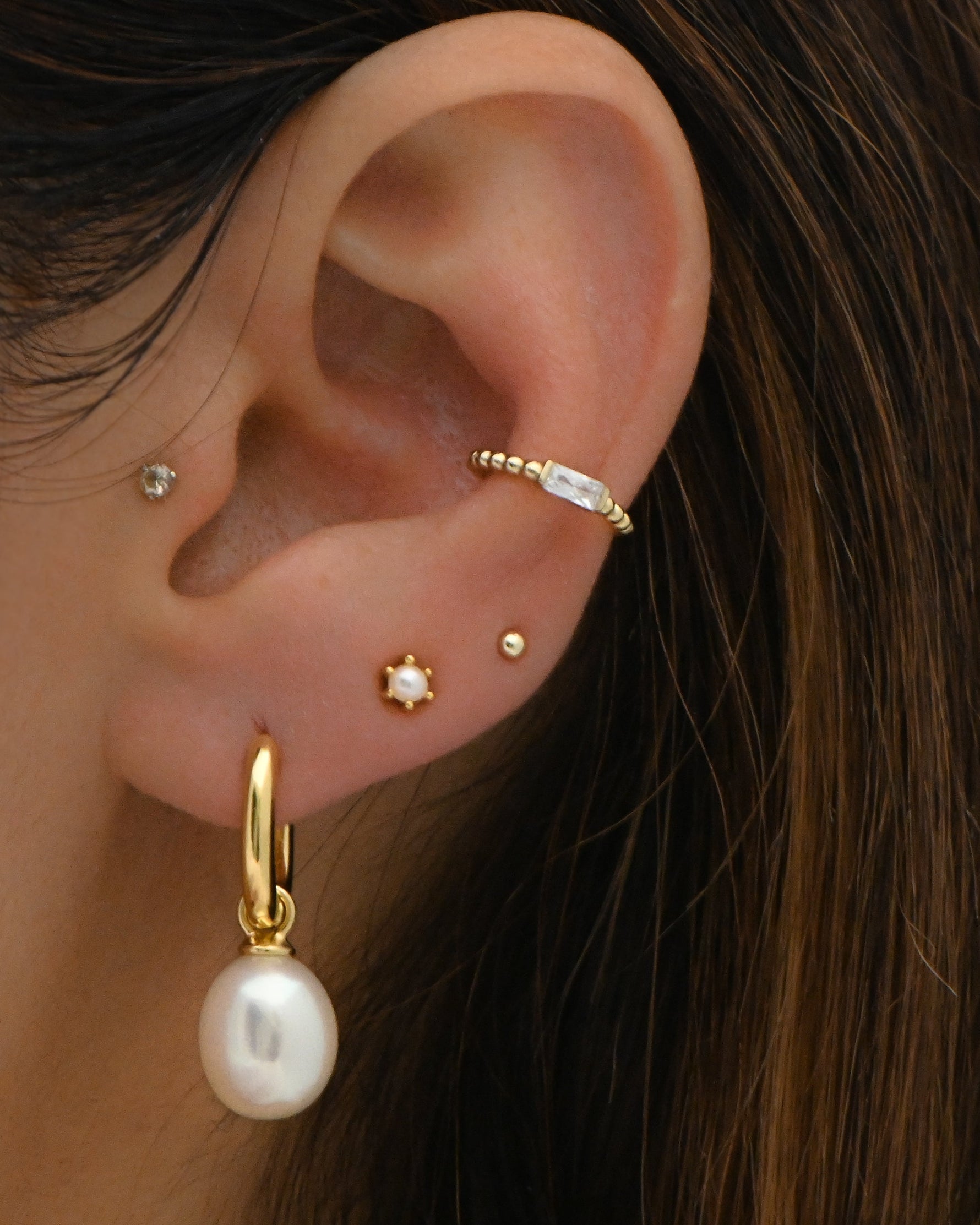 Model wearing gold pearl charm earrings, dainty pearl studs, ball studs, and a cz baguette ear cuff. 
