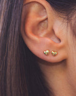 Model wearing two tiny gold plain heart stud earrings in a stack.