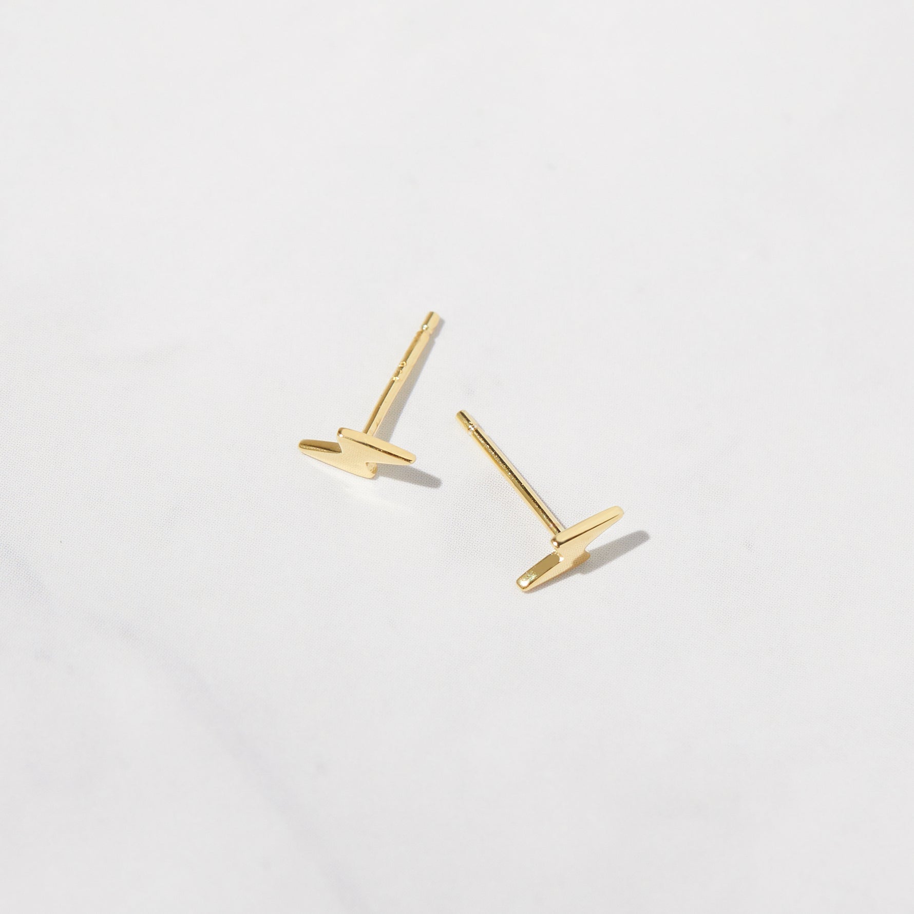 Delicate gold plain lightning bolt stud earrings on white marble background. 