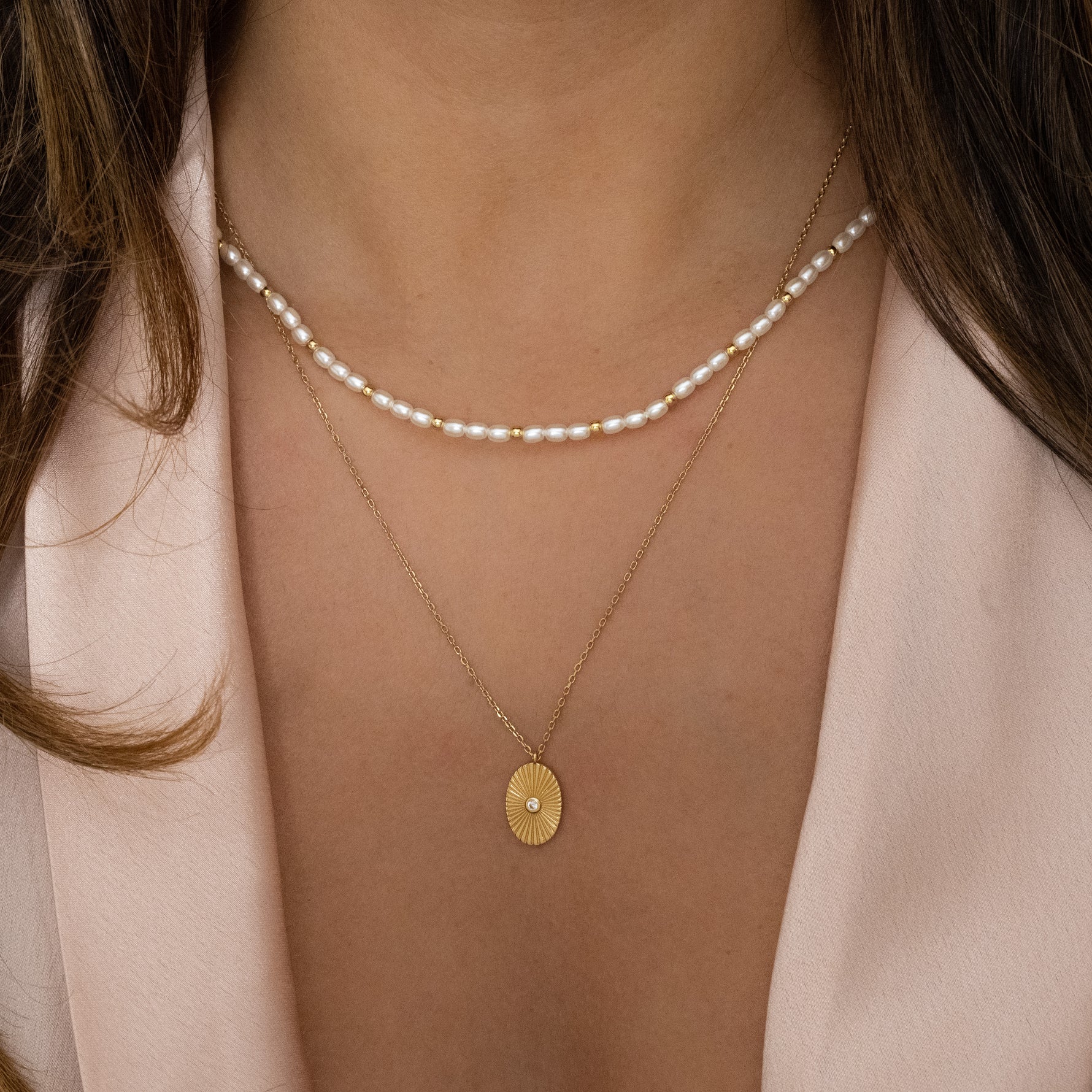 Model wearing gold pearl beaded necklace layered with a gold oval medallion necklace.