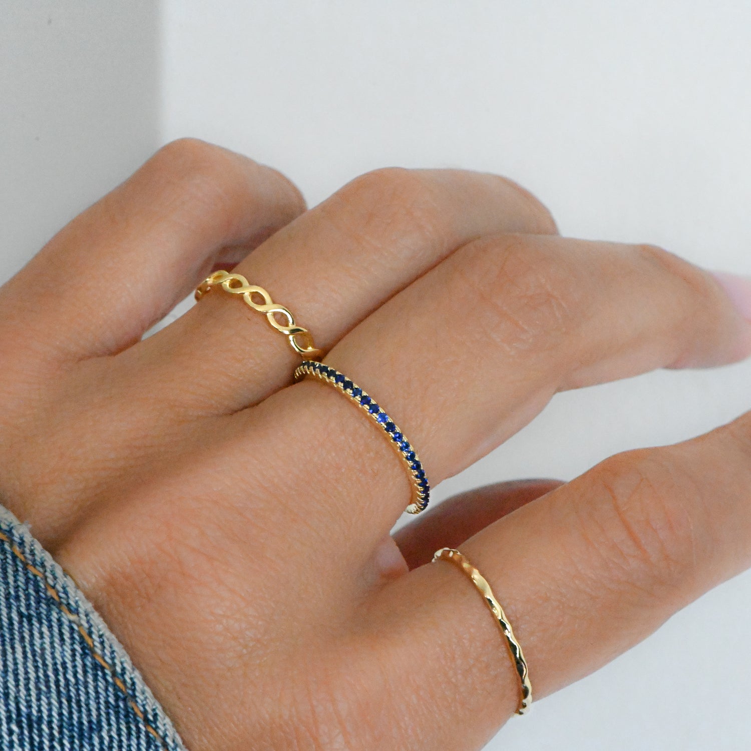 Model wearing gold ring stack featuring an open infinity band, a sapphire eternity ring, and a hammered thin band. 