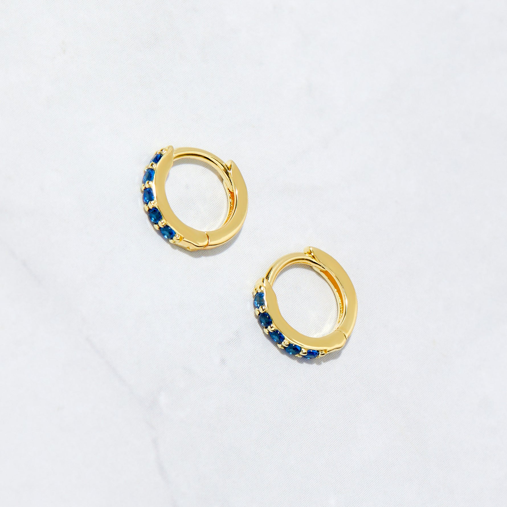 Gold sapphire birthstone huggie hoop earrings on white marble background.