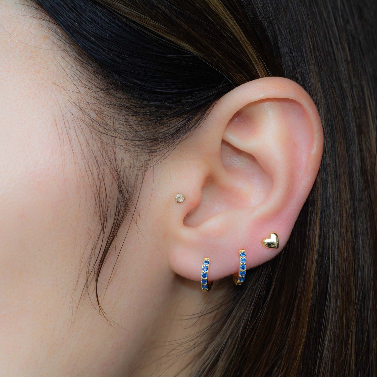 Model wearing three gold earrings in a stack featuring gold sapphire huggies and a heart stud earring. 