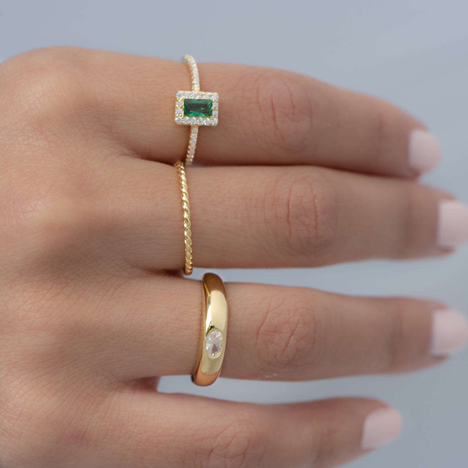 Model wearing gold ring stack including an emerald ring, a thin spiral band, and a bezeled dome ring. 