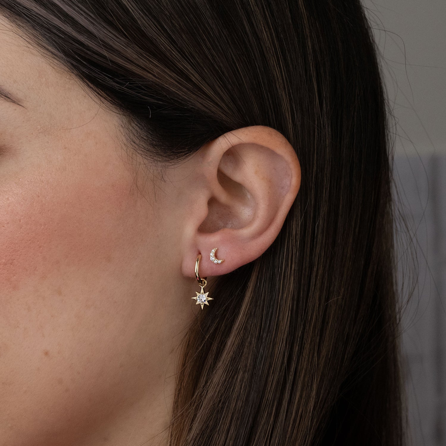 Model wearing dainty gold huggie hoop earrings with a sunburst cz charm dangling and a moon stud.