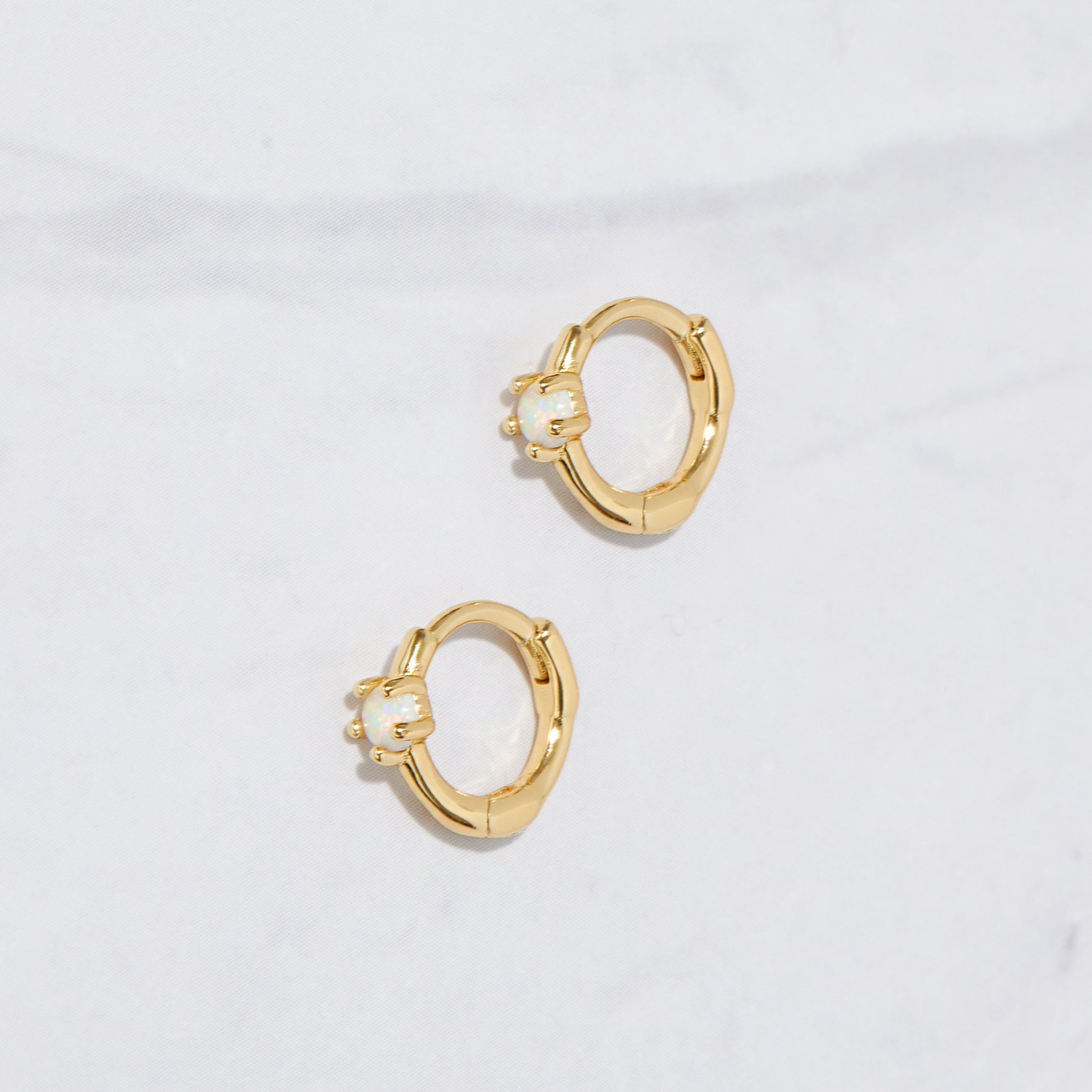 A closeup flat-lay picture of small gold hoop earrings. It features a solitaire opal stone.  