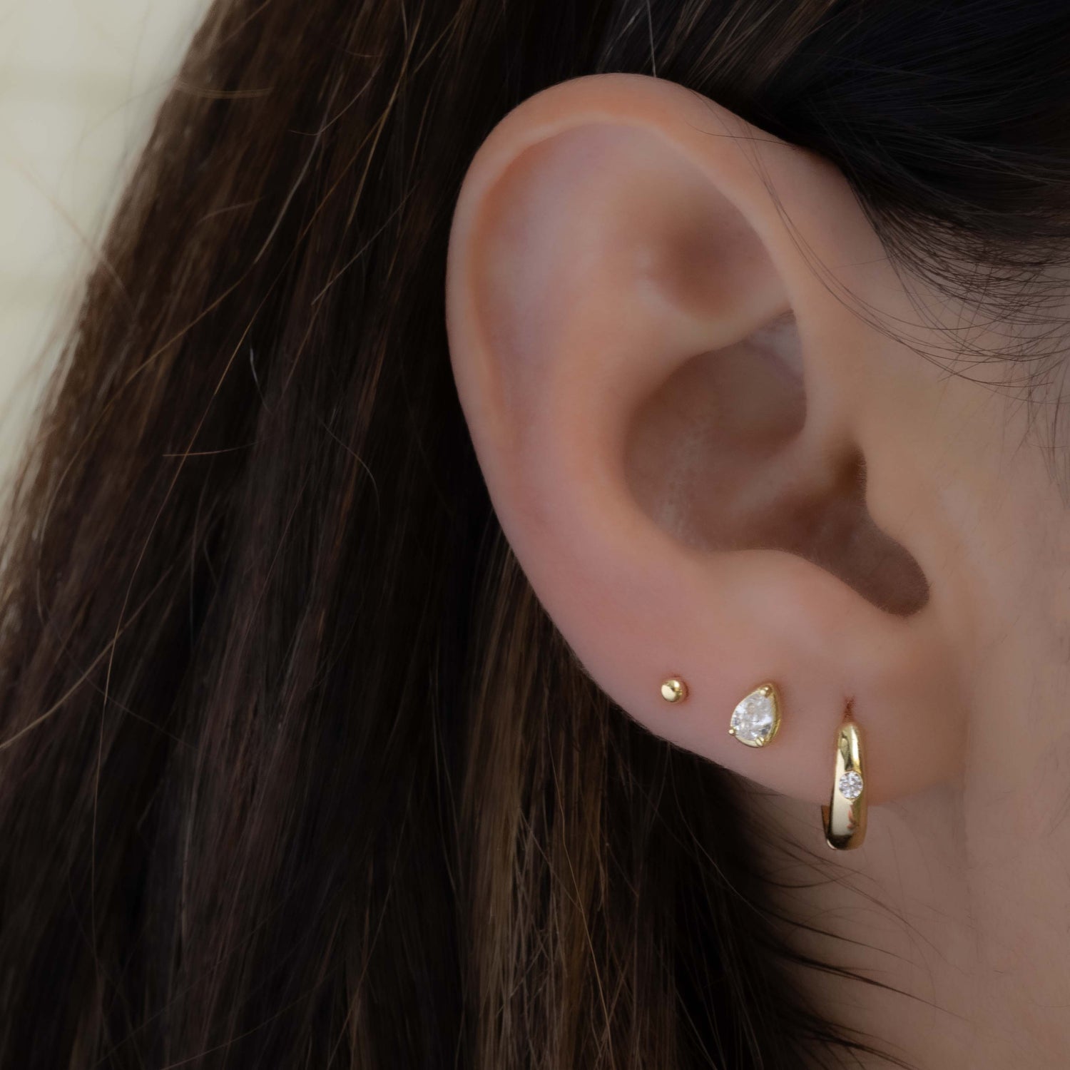 model wearing gold huggie hoop earrings, a dainty pear stud earring, and a small ball stud earring